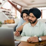 Cheerful young Indian woman cuddling and supporting serious husband working at home with laptop and counting on calculator